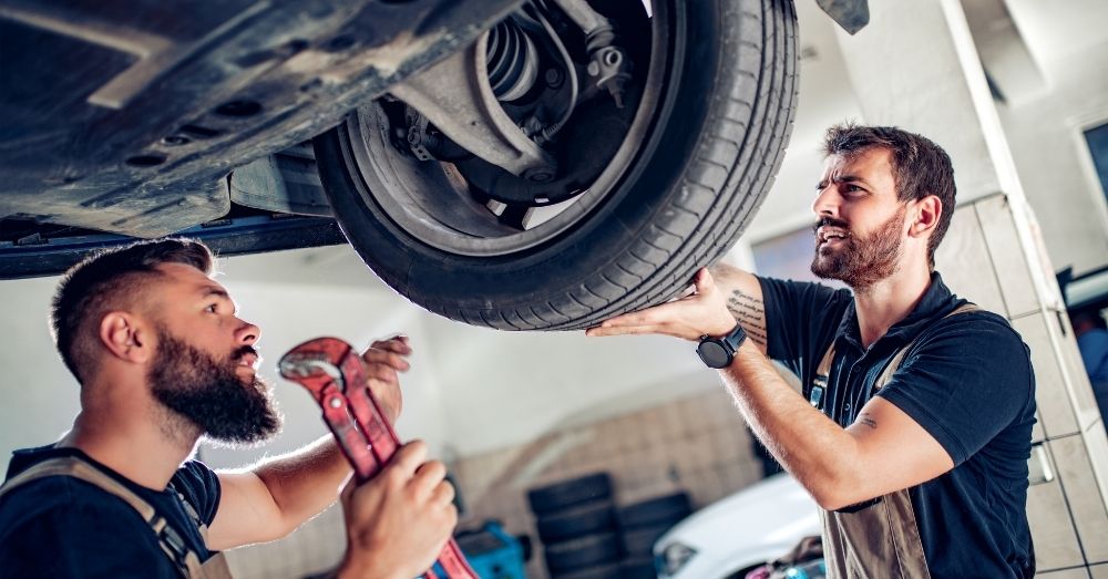 Two mechanics inspect a car's underside and tire.